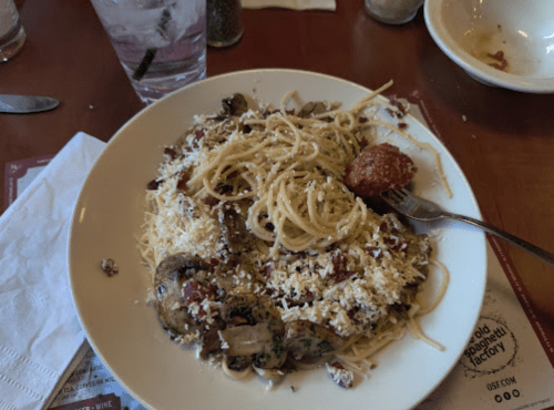 A plate of spaghetti topped with mushrooms, herbs, and a meatball, served with a glass of water in the background.