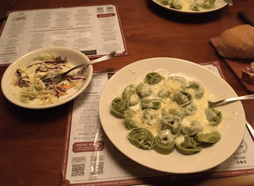 A plate of green tortellini in creamy sauce next to a small bowl of coleslaw on a wooden table with menus.