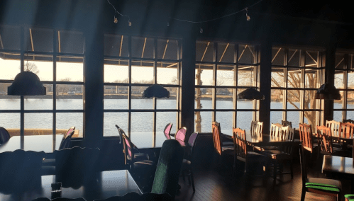 Interior of a restaurant with large windows overlooking a lake, featuring wooden tables and chairs in warm lighting.