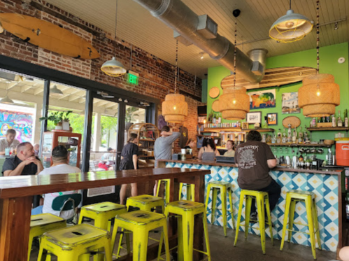 A vibrant café interior with colorful decor, bar stools, and patrons at the counter enjoying drinks and conversation.