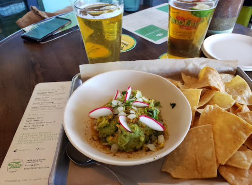 A bowl of guacamole topped with radishes and onions, served with tortilla chips and two glasses of beer.