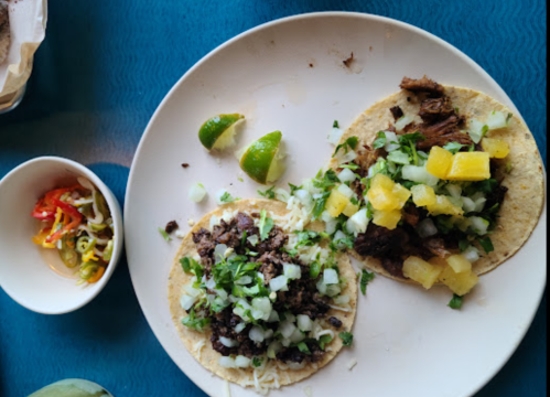 Two tacos on a plate, one with meat, cheese, and onions, the other topped with pineapple and cilantro, with lime wedges.