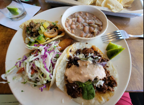 A plate with three tacos, beans, and a side of lime, featuring various toppings and fillings.