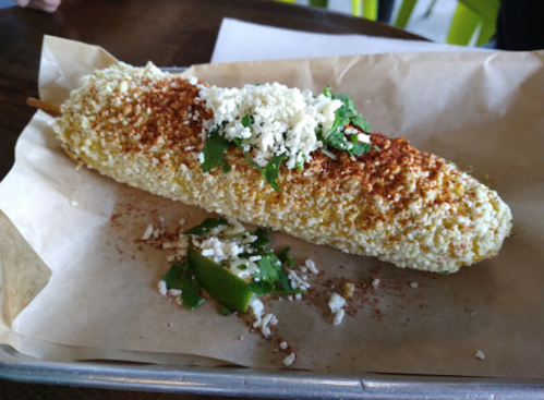 A corn on the cob topped with cheese, cilantro, and spices, served on a paper-lined tray with a lime wedge.