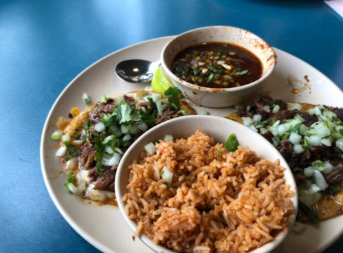 A plate of tacos topped with onions and cilantro, served with rice and a small bowl of sauce.
