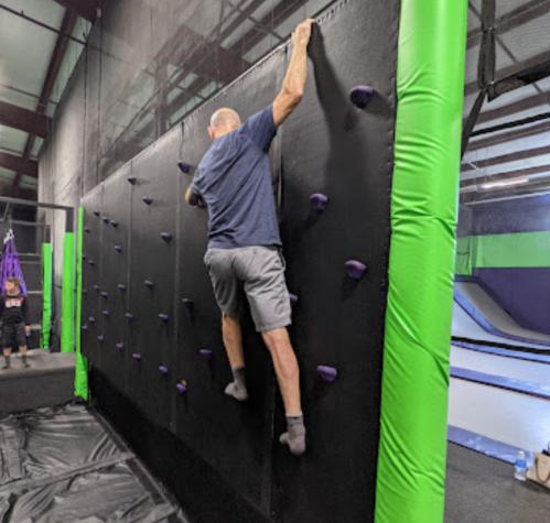 A person climbs a vertical wall with grips in a gym, while another person watches in the background.