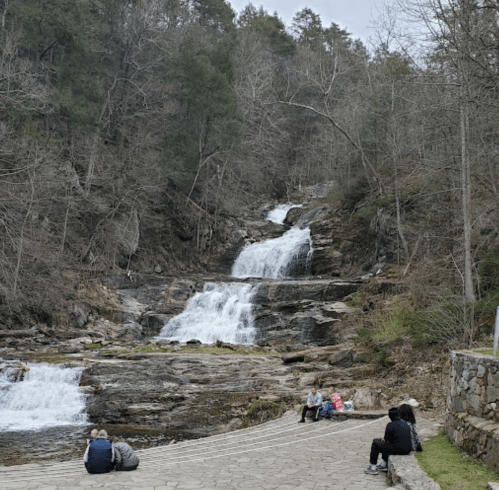 A serene waterfall cascades down rocky terrain, with people sitting on a stone path nearby, surrounded by trees.