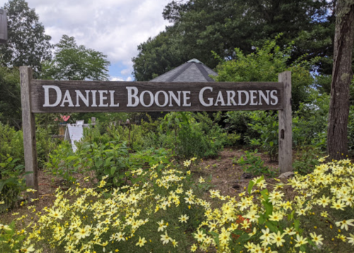 Sign for Daniel Boone Gardens surrounded by greenery and yellow flowers under a cloudy sky.