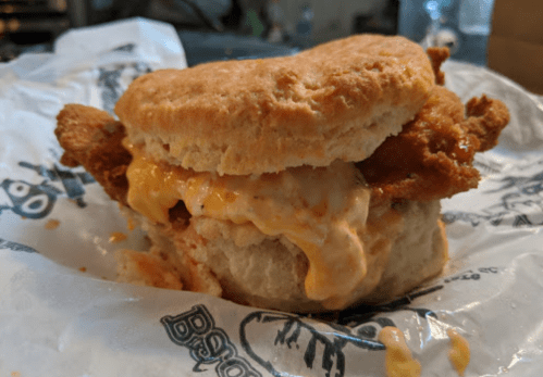 A close-up of a biscuit sandwich filled with fried chicken and melted cheese, wrapped in paper.