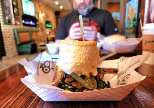 A large biscuit sandwich filled with greens and jalapeños, with a person using a phone in the background.