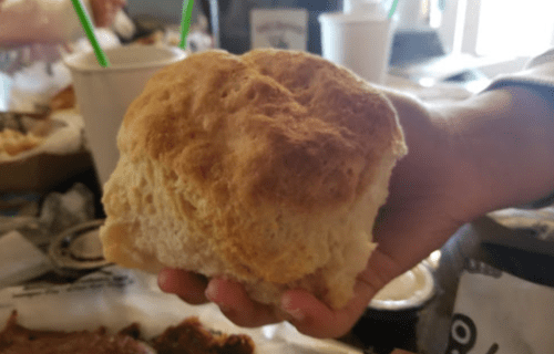 A hand holding a large, fluffy biscuit with a golden-brown top, surrounded by food and drinks on a table.