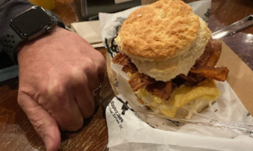 A hand holding a biscuit sandwich filled with bacon and cheese, resting on a paper wrapper on a wooden table.