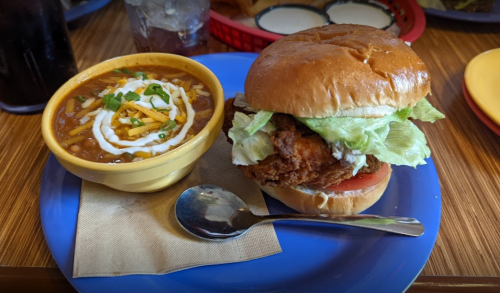 A plate with a crispy chicken sandwich topped with lettuce and tomato, alongside a bowl of chili with cheese and sour cream.