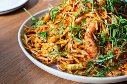 A plate of spicy pasta topped with fresh greens and shrimp, served on a wooden table.