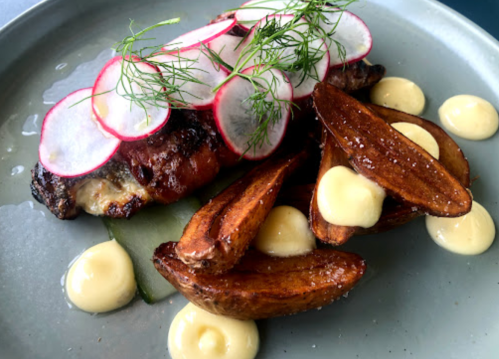 A plate featuring grilled meat topped with radish slices, alongside crispy potato wedges and dollops of creamy sauce.