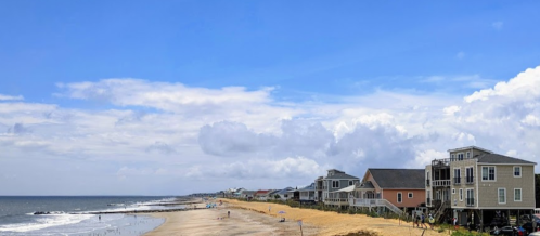 A scenic beach view with houses along the shore under a blue sky with scattered clouds.