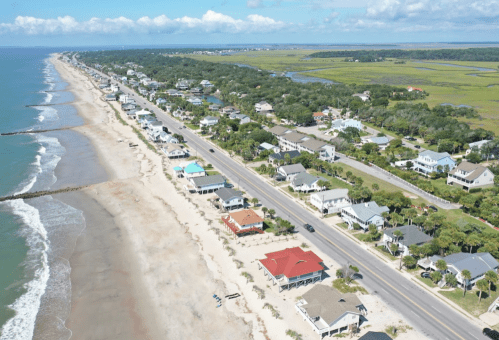 Aerial view of a coastal town with sandy beach, houses lining the shore, and lush greenery in the background.