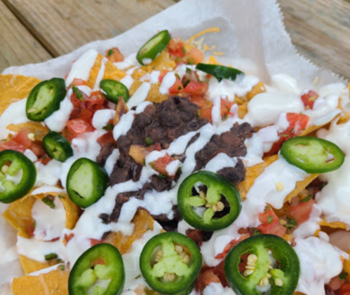 A close-up of nachos topped with cheese, black beans, diced tomatoes, jalapeños, and sour cream on a paper tray.