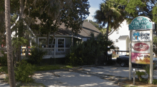 A quaint building with a porch, surrounded by trees, featuring a sign for Edisto Village Shops and nearby eateries.