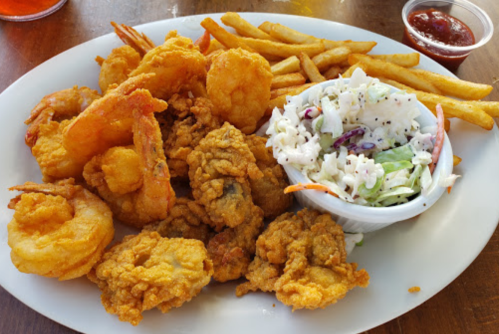A plate of fried seafood including shrimp, fish, and oysters, served with fries and coleslaw.