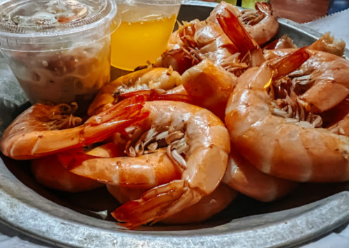 A platter of large shrimp with a side of sauce and a drink in clear cups.