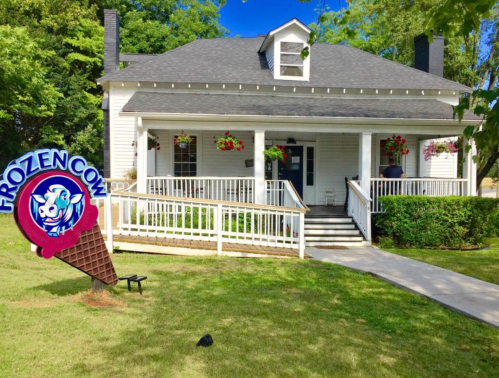 A charming white house with a porch, featuring a colorful "Frozen Cow" ice cream sign and flower baskets.