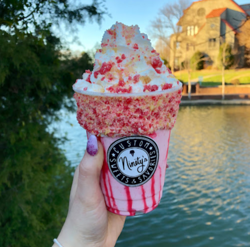 A hand holding a colorful milkshake topped with whipped cream and crushed toppings, with a scenic waterfront in the background.