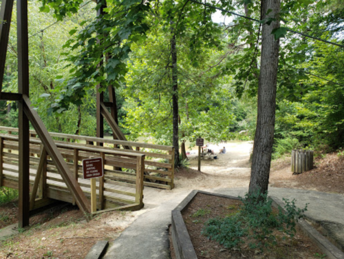 A wooden bridge leads to a sandy area surrounded by trees, with picnic spots visible in the background.