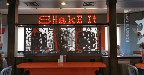 A restaurant interior featuring a bright "Shake It" sign and a decorative screen with a pixelated design.