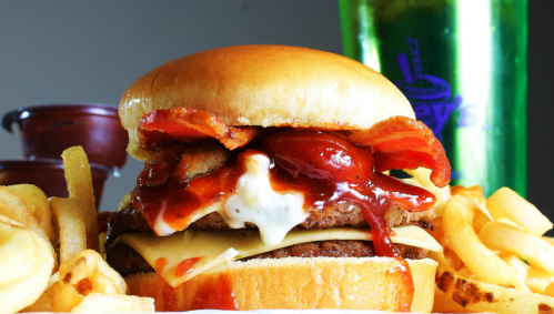 A close-up of a juicy burger with bacon, cheese, and sauces, served with fries and a drink in the background.