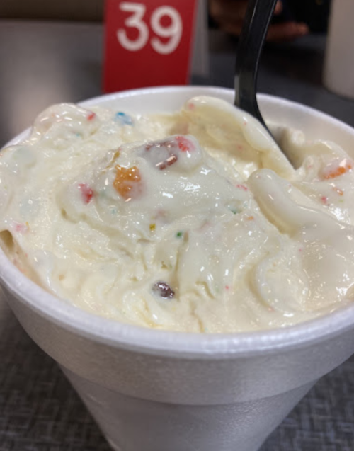 A close-up of a cup of creamy ice cream with colorful sprinkles, served with a black spoon.