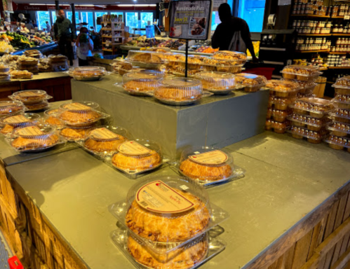 A display of various baked pies in clear containers at a market, with shoppers in the background.