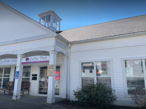 Exterior of a white building with a sign for "Cutie Pies" and a small tower, featuring a bench and accessible parking sign.