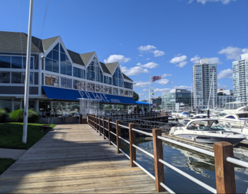 A waterfront restaurant with a blue awning, boats docked nearby, and modern buildings in the background under a clear sky.