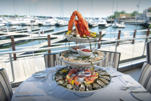 A tiered seafood platter featuring shrimp, oysters, and crab, set against a marina view with boats in the background.
