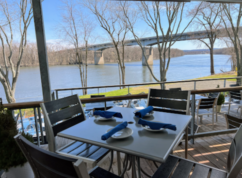 A riverside dining area with a table set for four, overlooking a bridge and calm water surrounded by bare trees.