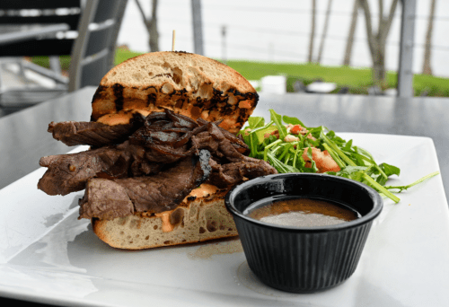 A grilled steak sandwich on toasted bread with sautéed onions, served with a side salad and dipping sauce.