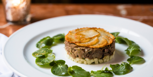 A gourmet dish featuring layered meat, crispy potatoes, and fresh greens arranged on a white plate.