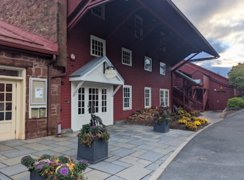 Red building with white trim, large windows, and flower planters, set against a cloudy sky. Pathway leads to entrance.