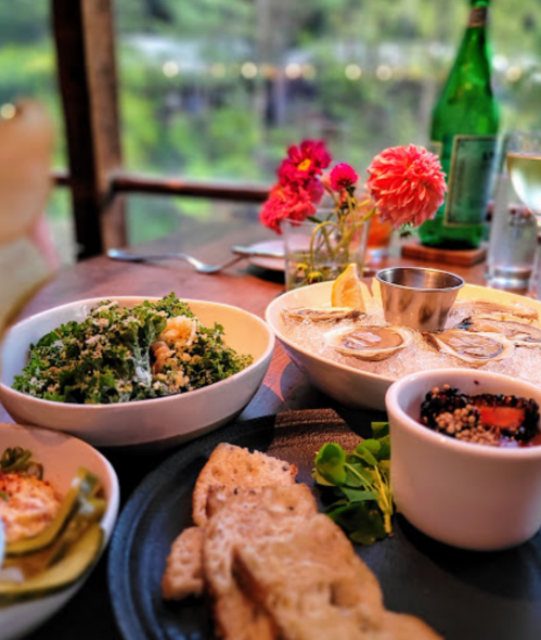 A table setting with a salad, oysters, bread, and flowers, with a scenic view in the background.