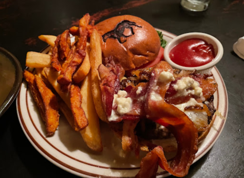 A plate with a burger topped with bacon and sauce, served with regular and sweet potato fries and a side of ketchup.