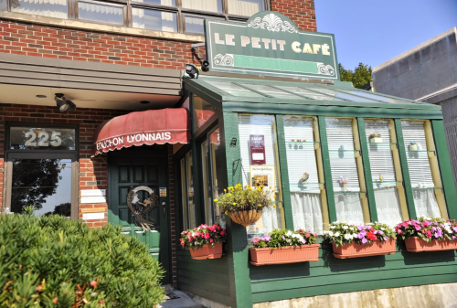 A charming café with a green glass entrance, flower boxes, and a red awning, set in a brick building.