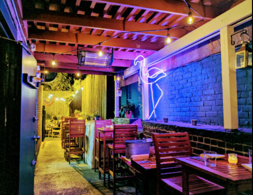 Cozy outdoor dining area with wooden tables, string lights, and a neon flamingo against a blue wall.
