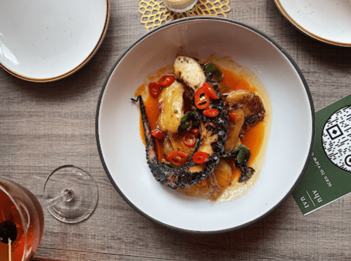 A white bowl filled with grilled octopus, potatoes, and sliced red chilies in a flavorful sauce, set on a wooden table.
