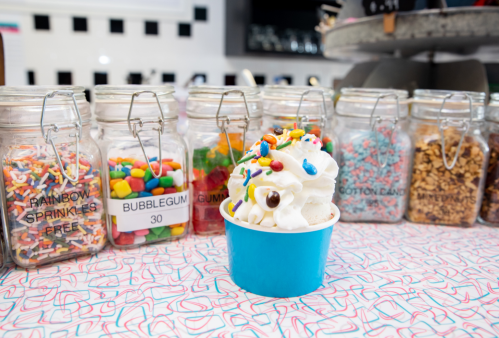 A blue cup filled with soft serve ice cream topped with colorful sprinkles, in front of jars of various toppings.