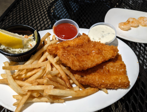 A plate of crispy fried fish fillets with fries, coleslaw, and dipping sauces, alongside a small plate of shrimp.