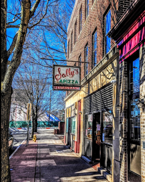 A street view of Sally's Pizza restaurant with a sign, surrounded by brick buildings and bare trees.