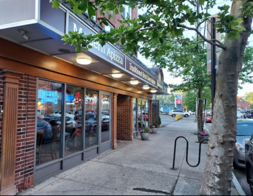 A street view of a pizza restaurant with outdoor seating, trees, and neon signs in the evening light.