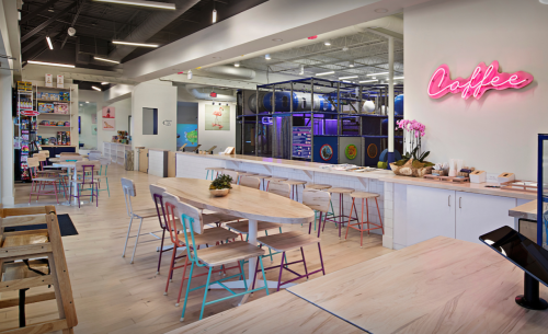 Bright café interior with wooden tables, colorful chairs, and a neon "Coffee" sign. Shelves filled with products in the background.