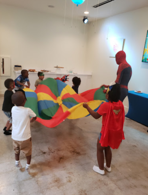 Children playing with a colorful parachute while a person in a superhero costume supervises the activity.